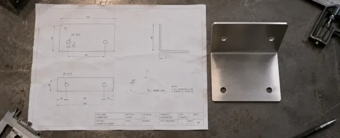 A printed 2D engineering drawing of a sheet metal bracket placed next to the fabricated metal part on a workbench
