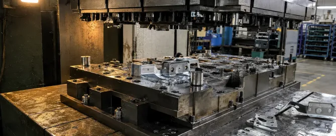 A progressive stamping die mounted on a hydraulic press bed during tryout phase with sample automotive bracket parts on the work surface